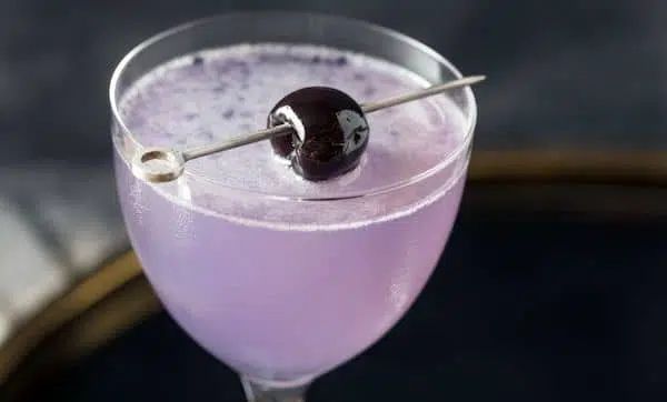 This is a lavender colored sambuca cocktail garnished with a cherry on top. 